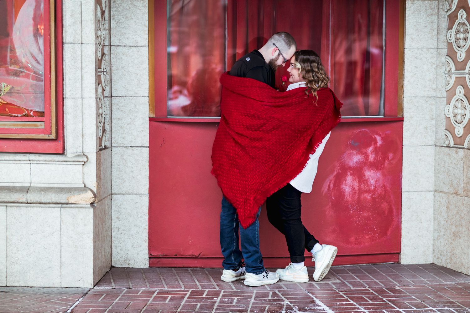 A couple wrapped in a red blanket share a tender moment together against a red door.
