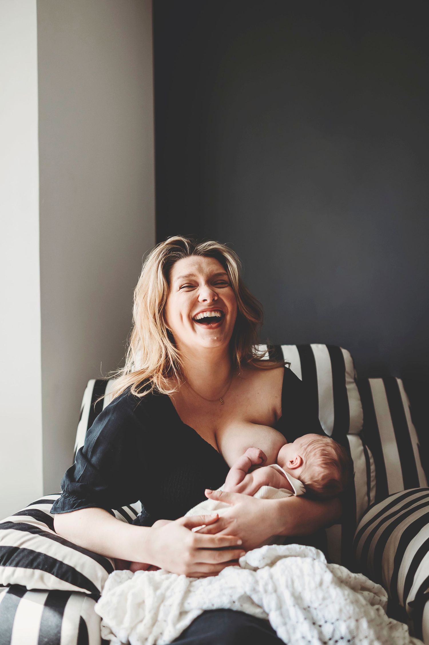 A joyful moment shared on a striped couch as a mother cradles her sleeping newborn, laughing with pure happiness.