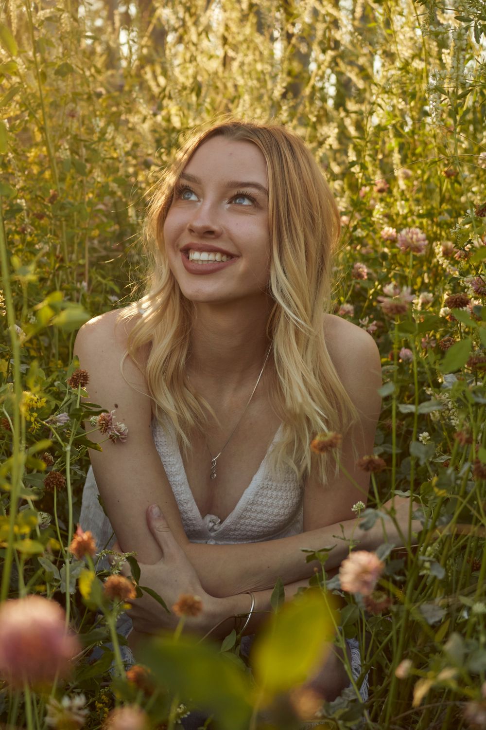 Young woman sitting in a field surrounded by flowers. She has curled hair and is wearing jeans and a tank top