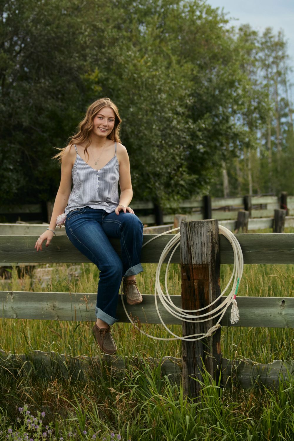 young woman sitting on a wooden fence in jeans and tank top, smiling in a rural field with flowers and a rope, summer countryside vibe.