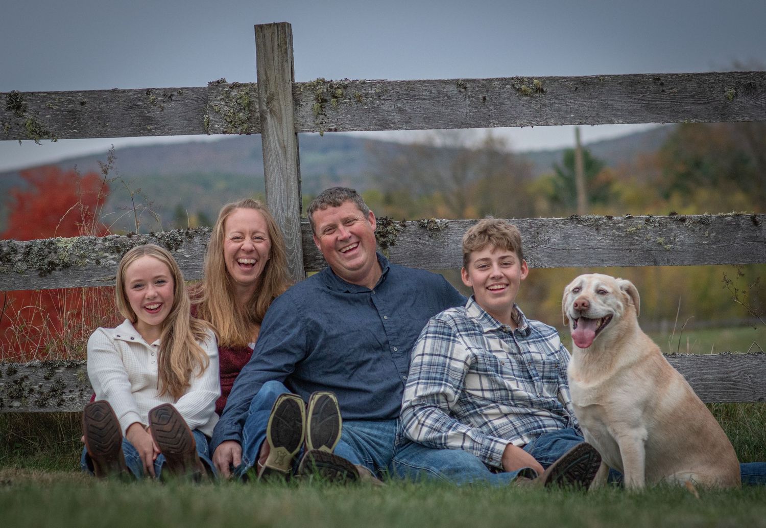 Happy family of four sitting on grass with yellow lab dog by wooden fence in autumn countryside. Photo by Xenophon Photography in Maine.