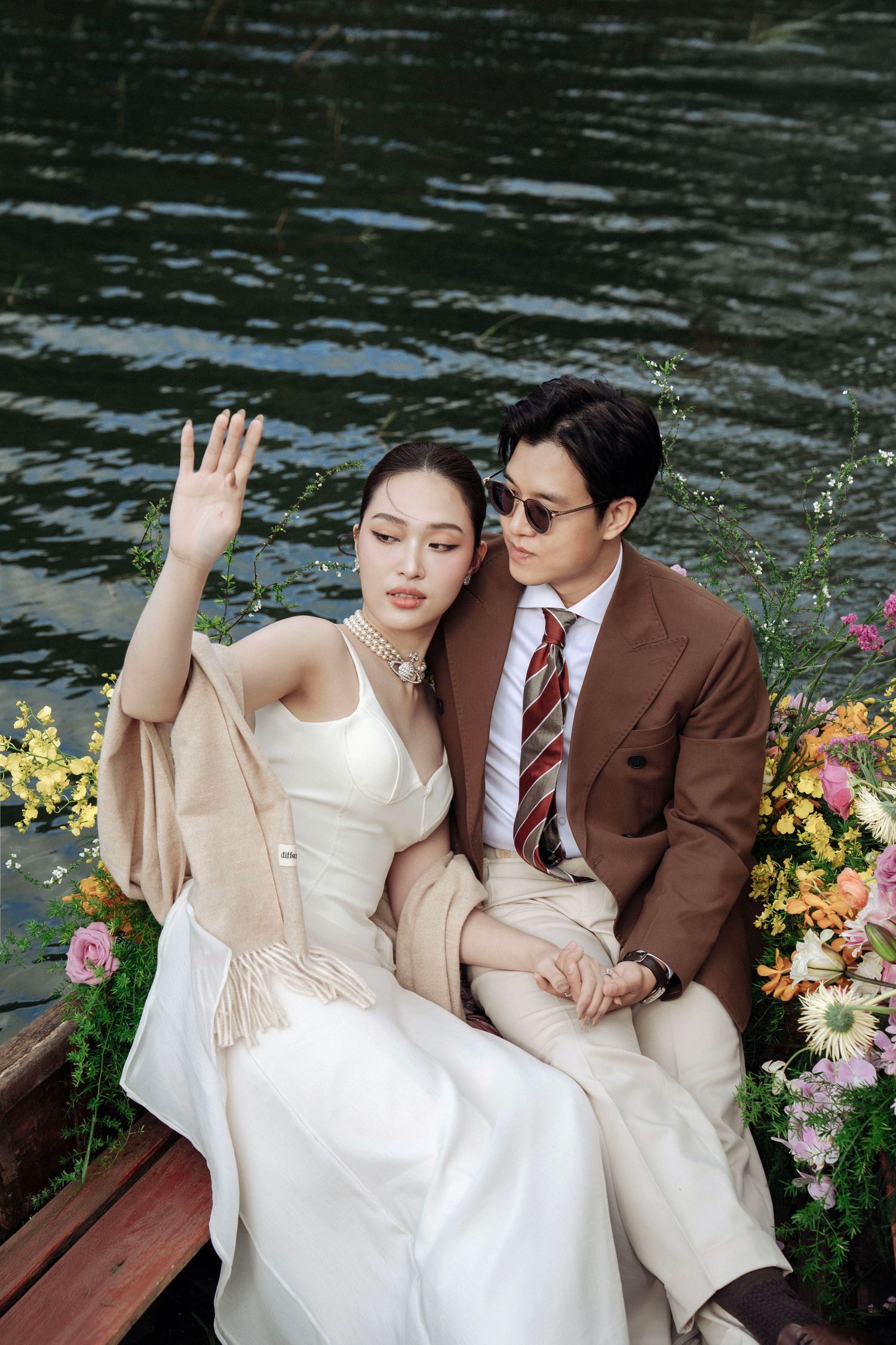 Bride in white dress with beige shawl leans on groom in brown suit beside colorful flowers on boat.