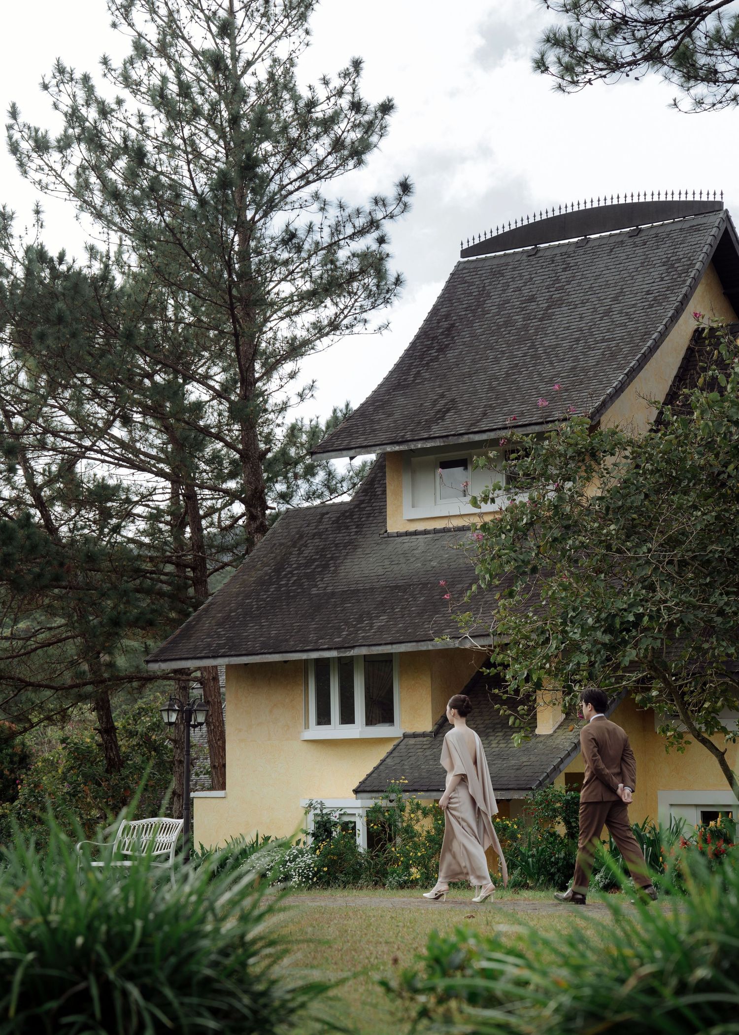 Couple walks through garden of charming yellow French colonial villa surrounded by pine trees.