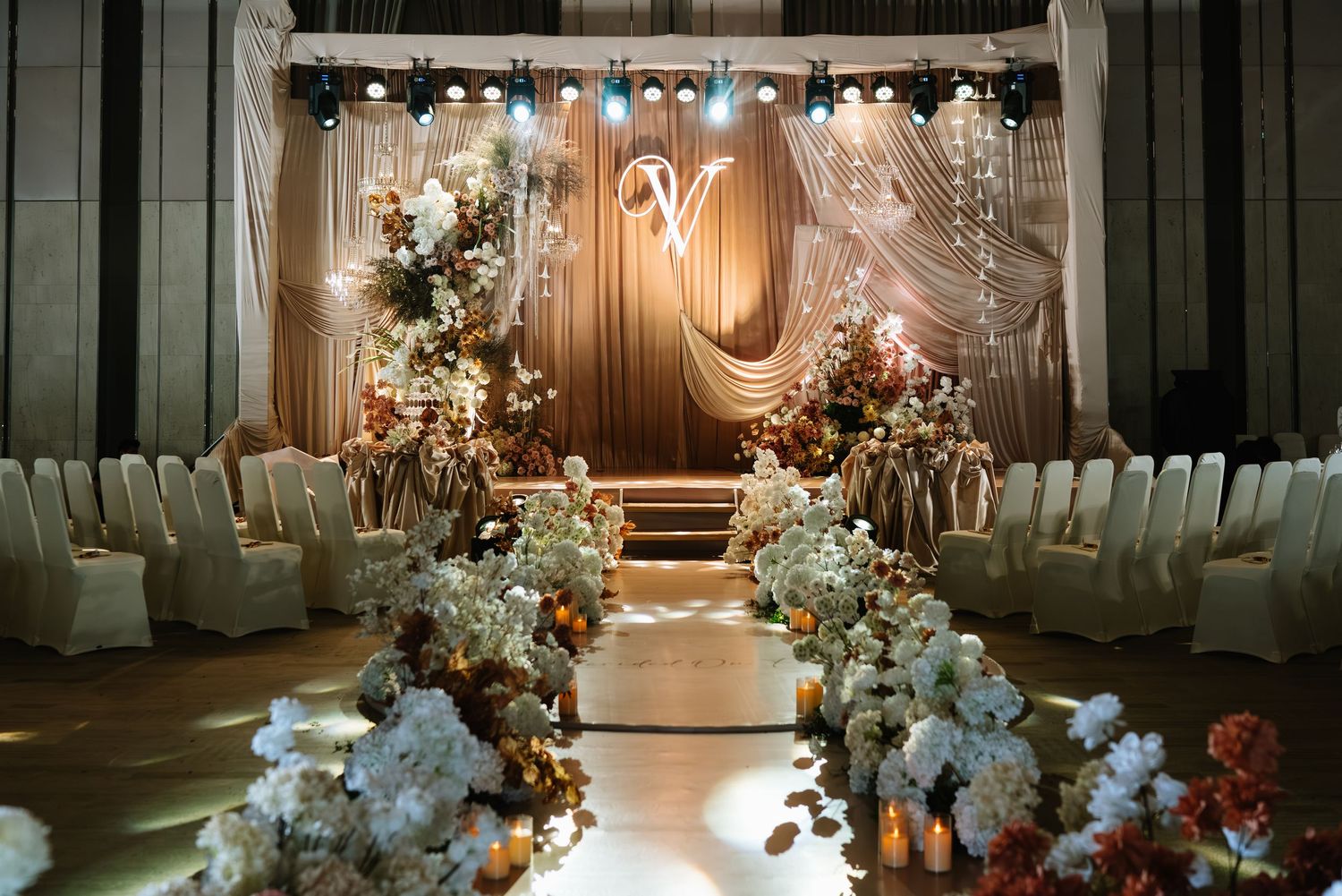 Sophisticated wedding ceremony hall with floral aisle arrangements, draped backdrop, and warm stage lighting.