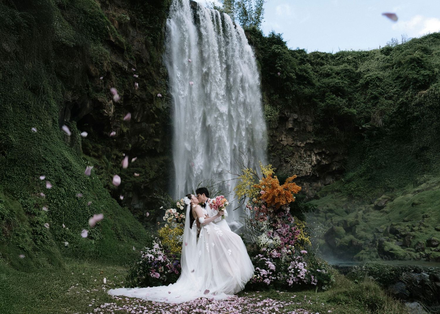 Bride and groom sharing romantic kiss in front of stunning waterfall surrounded by colorful flowers.