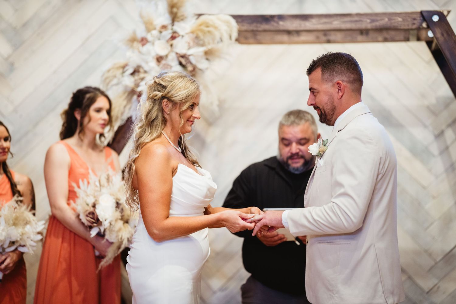 A couple exchanges rings during their wedding ceremony beneath a decorative arch with pampas grass.