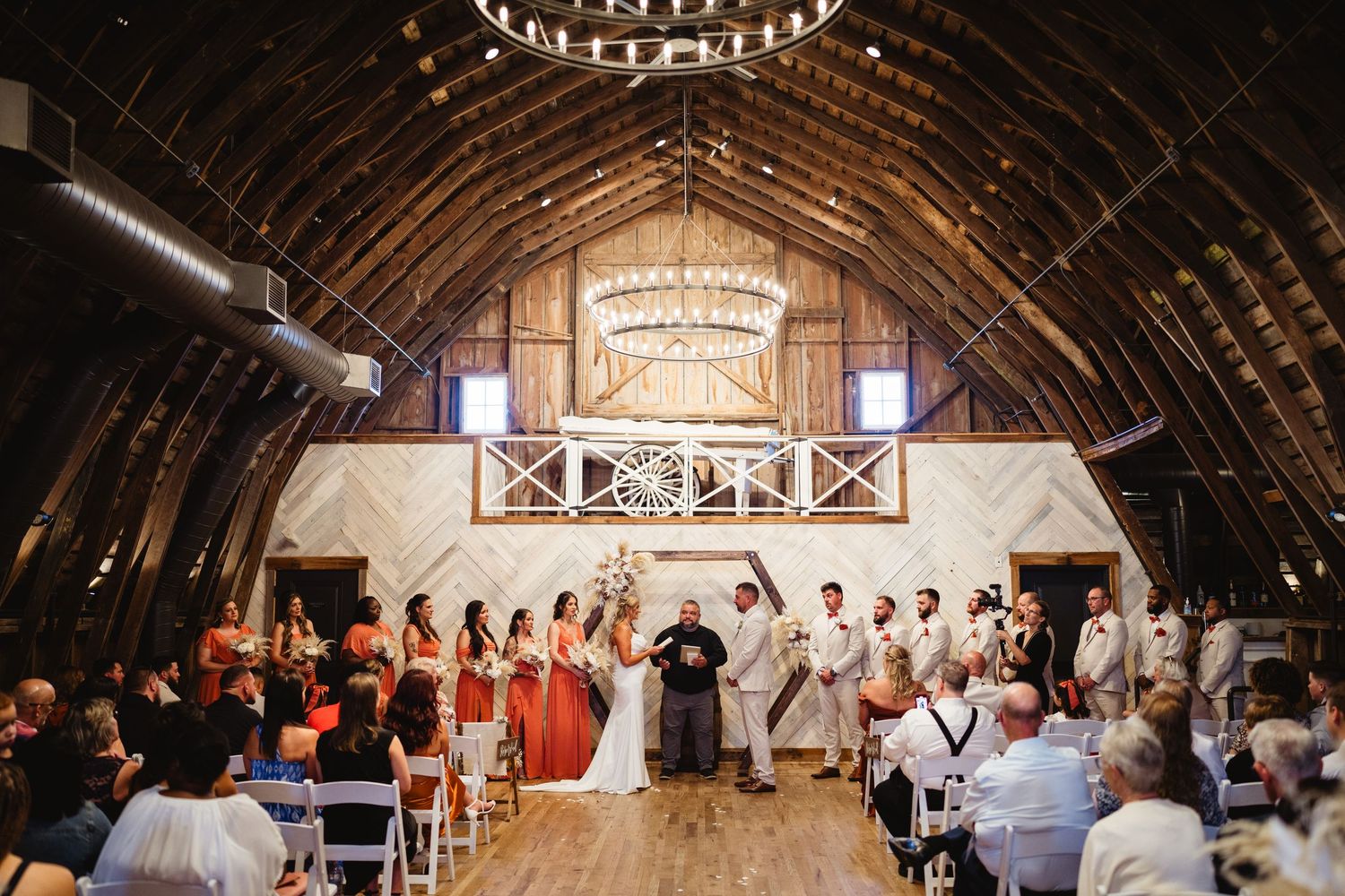 A rustic barn wedding ceremony with exposed wooden beams and chandeliers as guests witness the nuptials.