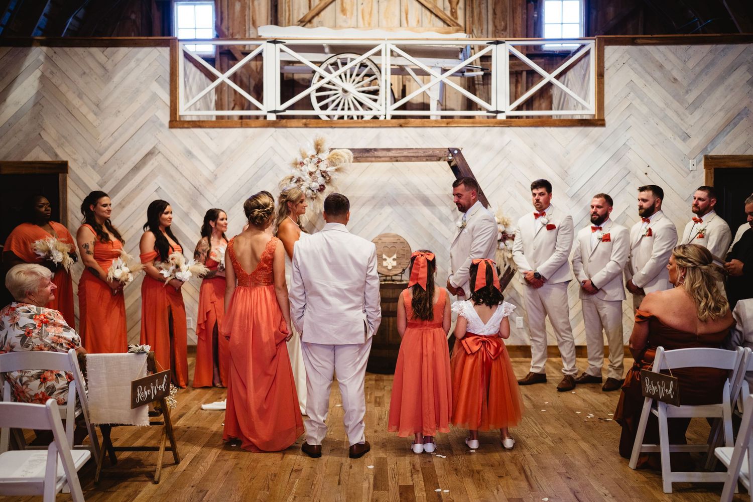 A wedding party stands in formal attire with bridesmaids in coral dresses and groomsmen in light suits at an indoor venue.