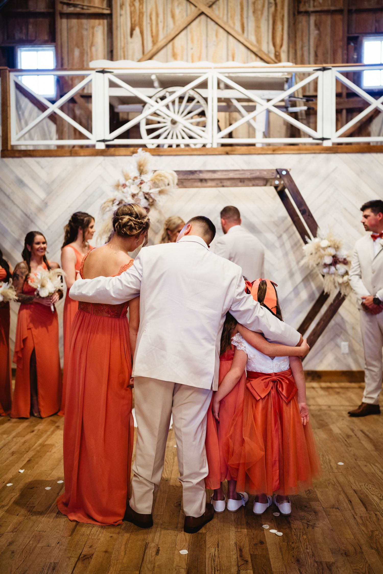 Wedding party members in white and coral attire share an embrace during a rustic barn reception celebration.