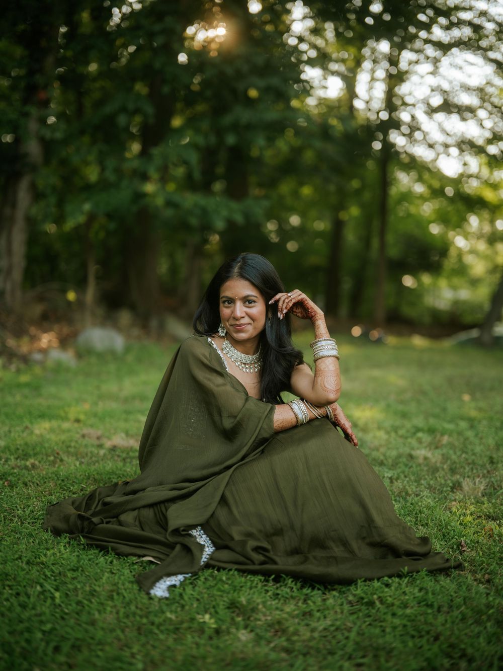 A person in a flowing olive green traditional dress sits gracefully on grass in a sunlit wooded setting.  Hilton Pearl River NY