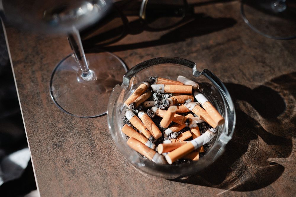 Glass ashtray filled with cigarette butts sits on a dark brown rustic surface in moody lighting.