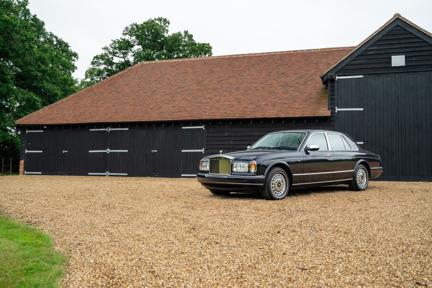 Classic car parked in front of rustic brown and black barns with gravel driveway.