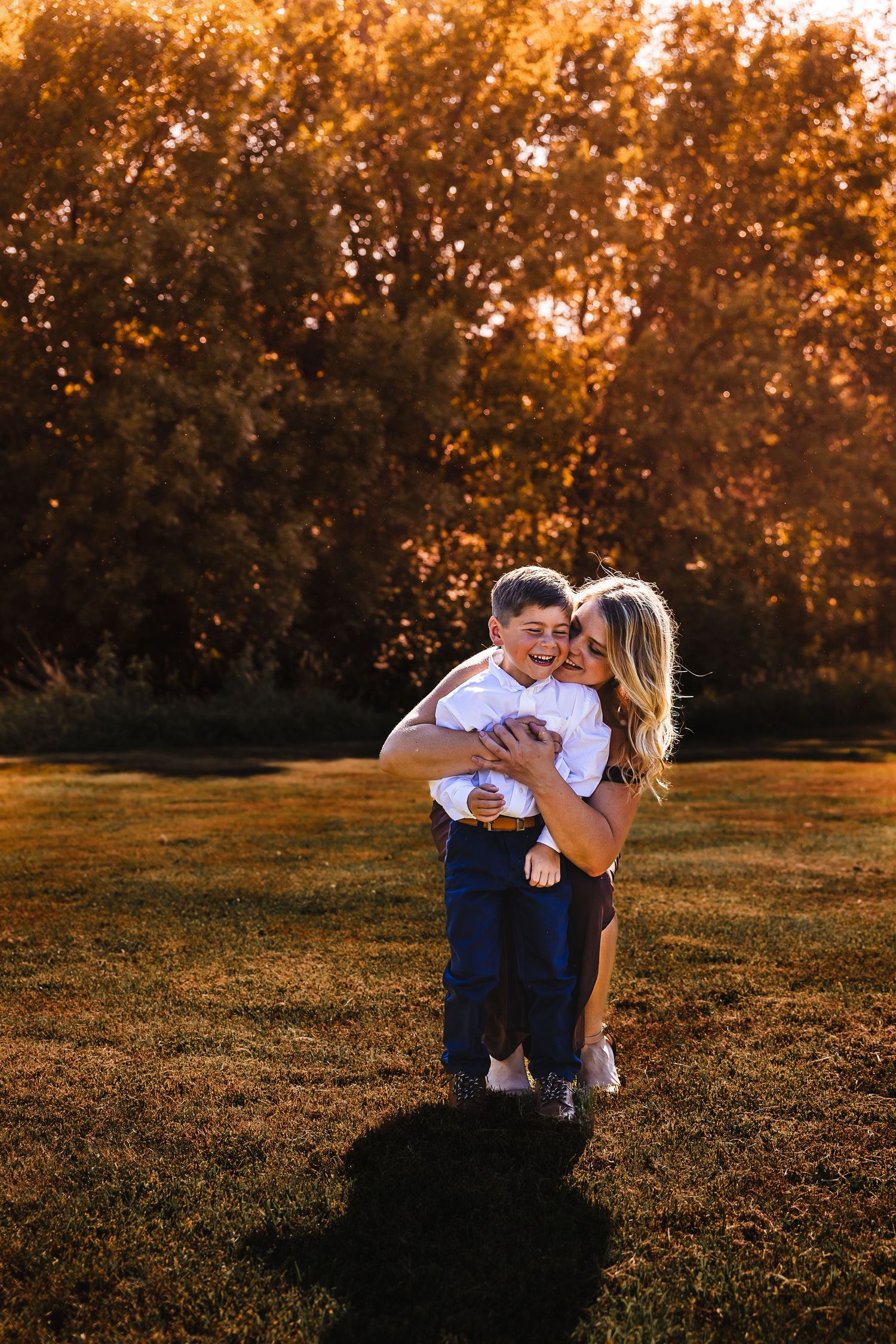 A couple embraces in a sunlit field during golden hour as autumn trees glow warmly in the background.