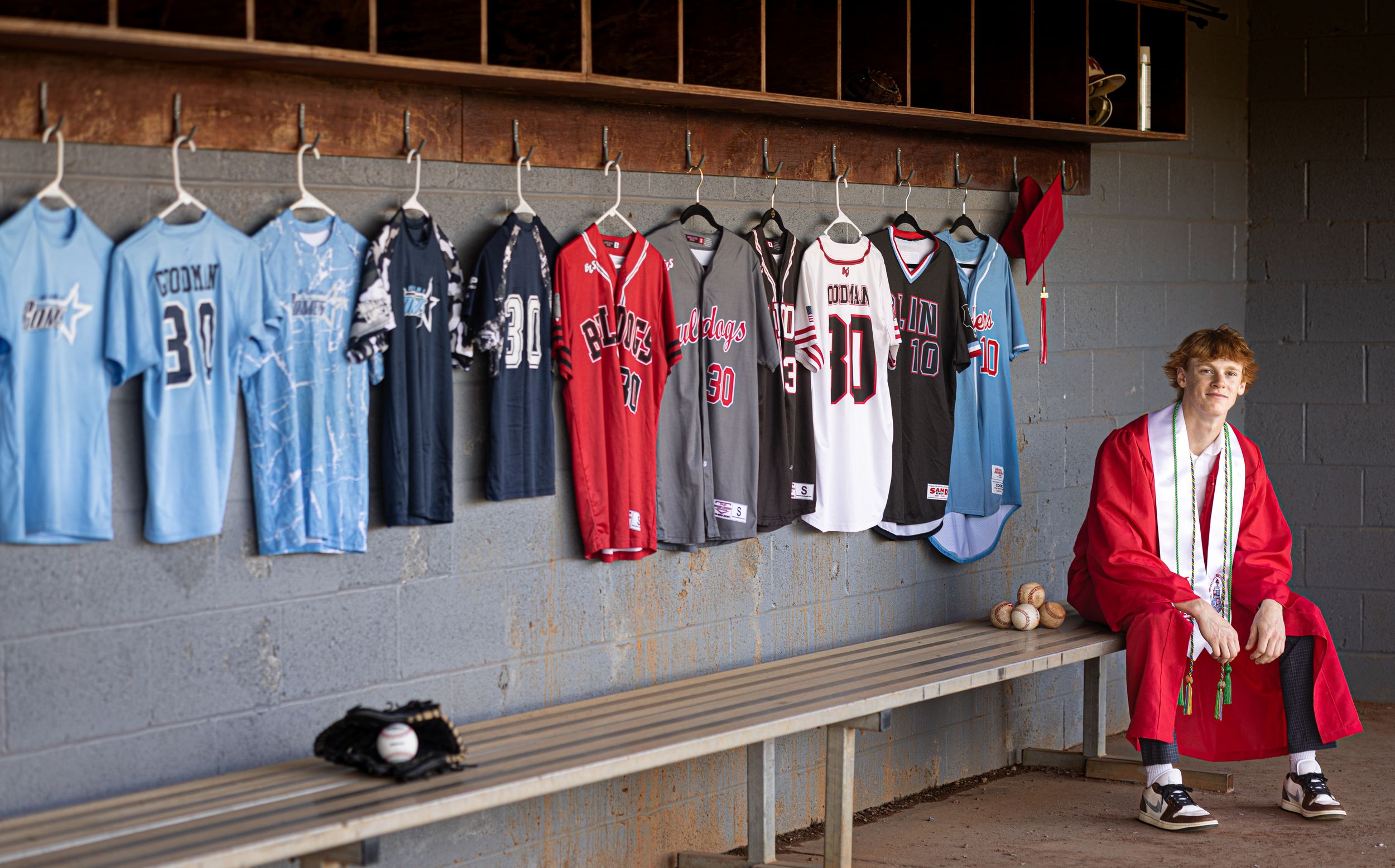 Baseball jerseys and uniforms hang on a locker room wall next to wooden bench and seating area.