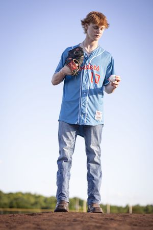 Baseball player in blue jersey and jeans stands on a dirt mound against a bright sky background.