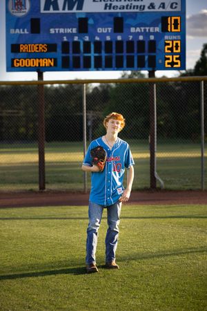 Little league baseball player stands in uniform on grass field at sunset with scoreboard visible in background.