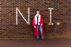 A graduate in red academic regalia poses against a brick wall during commencement ceremonies.