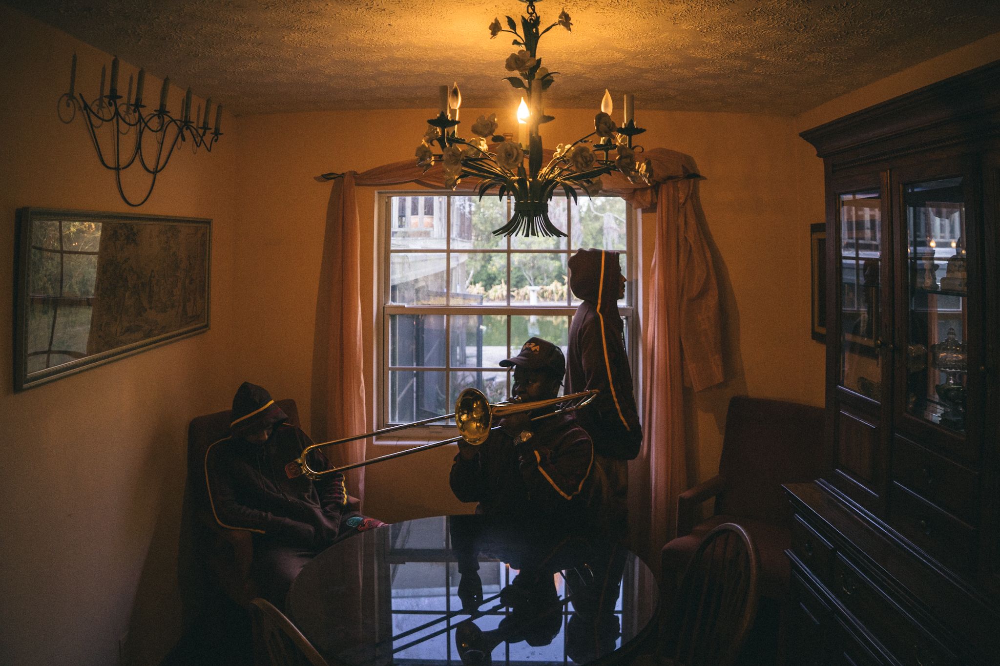 A classic piano sits in a dimly lit room with warm lighting from an ornate chandelier overhead and sunlight through windows.