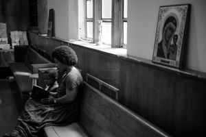 In a dimly lit church pew, someone sits praying in front of stained glass windows and an icon on the wall.
