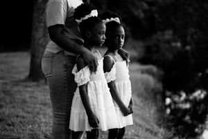 Two children in white dresses stand together outdoors while an adult gently touches their hair.