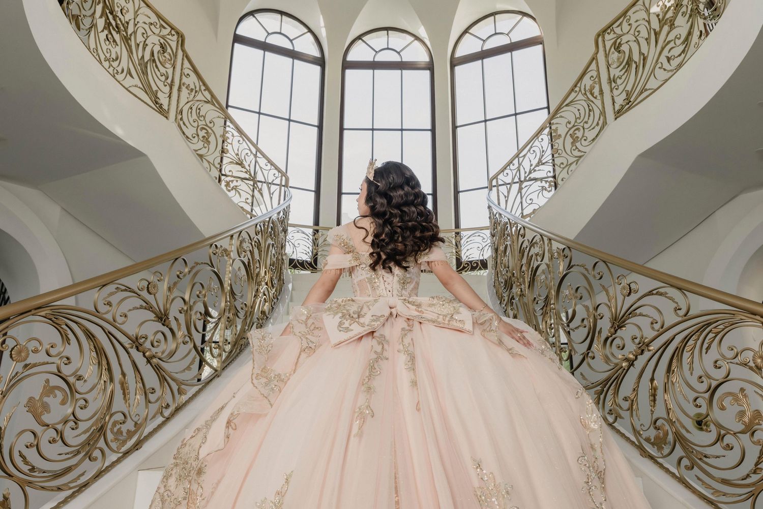Ornate staircase with elegant wrought iron railing curves upward in a grand entrance hall with arched windows.