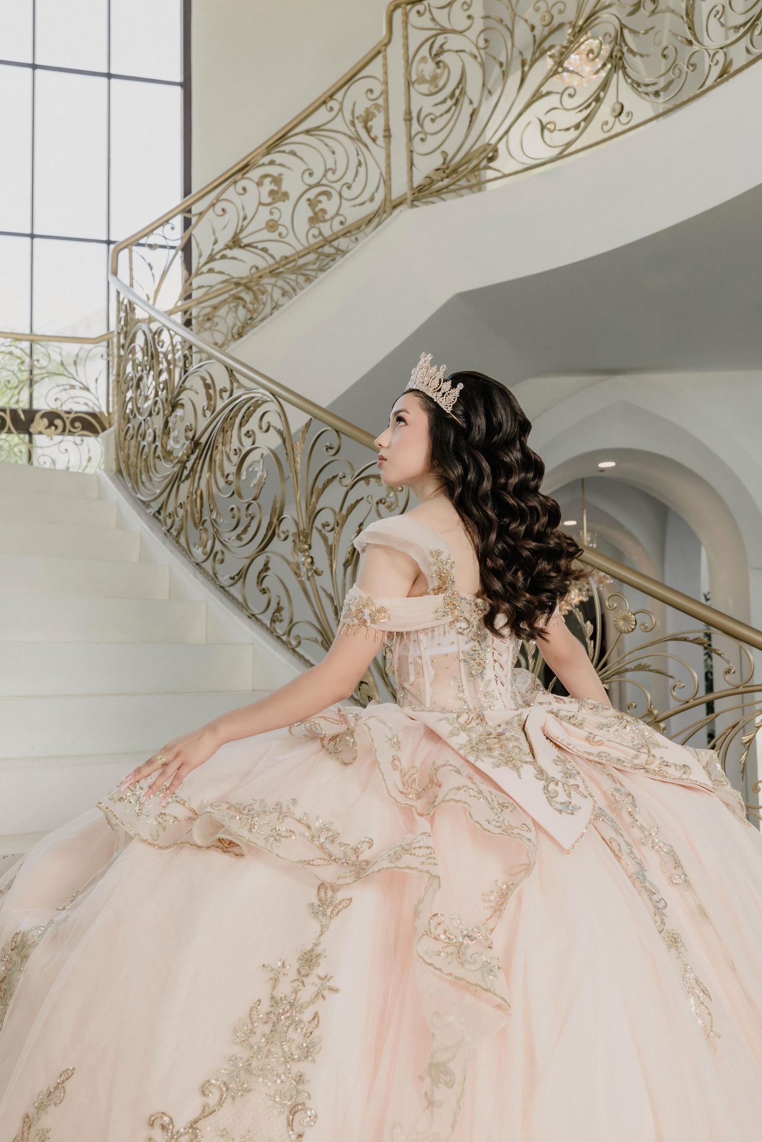 A flowing blush pink ball gown with gold embellishments cascades elegantly down an ornate white staircase.