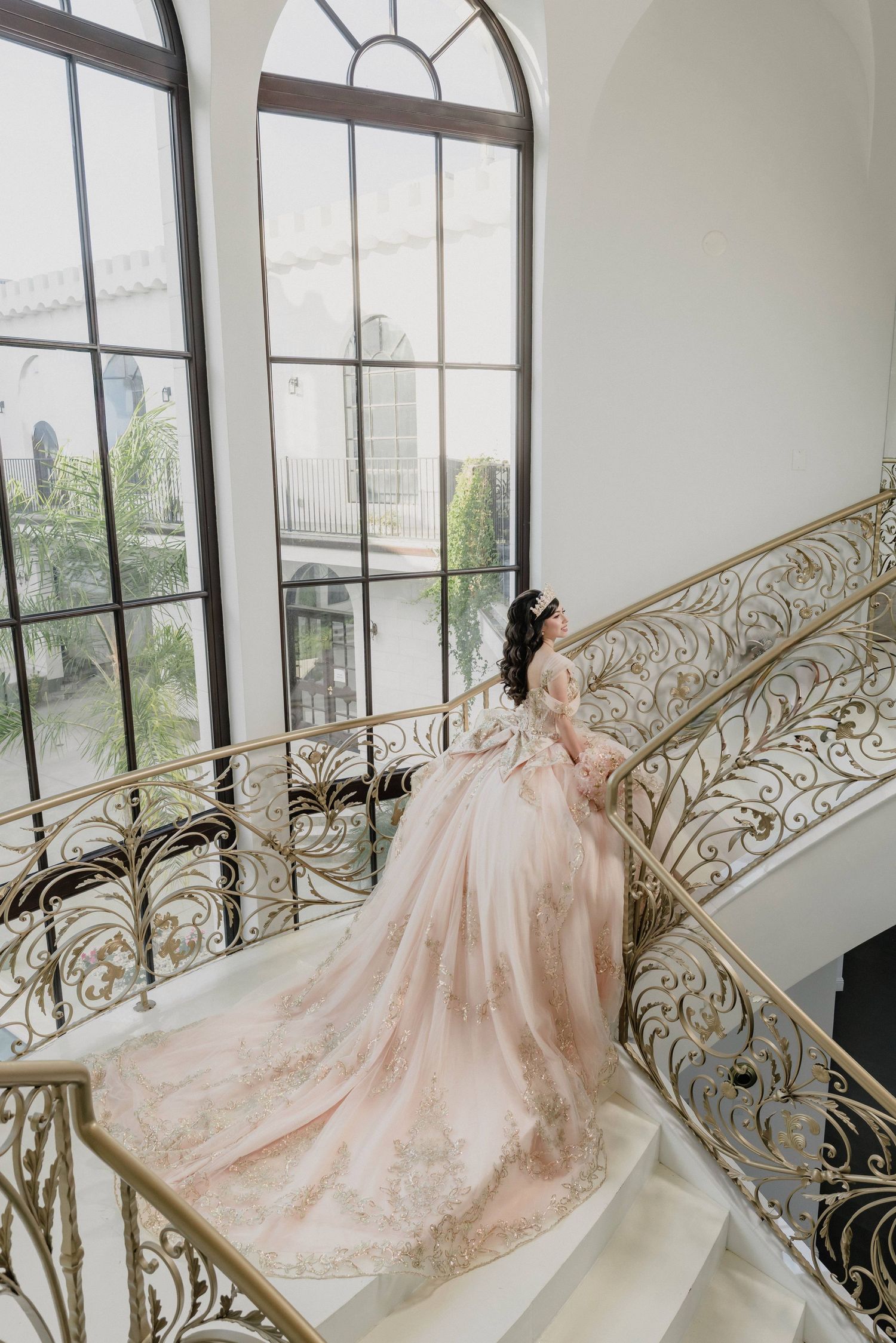 Elegant blush pink ballgown displayed against ornate gold railings and large windows in an indoor venue.