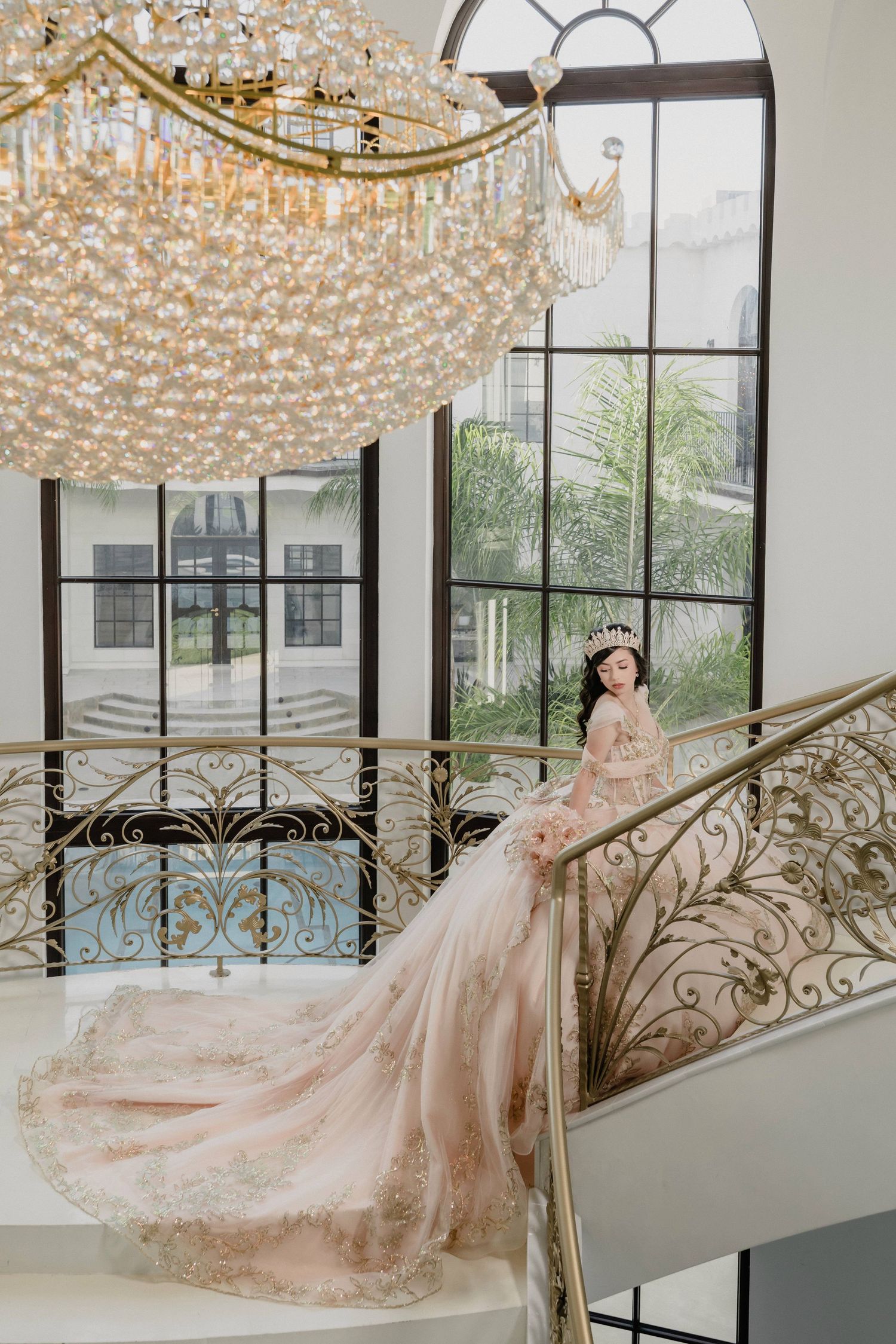 Luxurious ballgown photographed beneath crystal chandeliers in an elegant white interior with sweeping staircase.