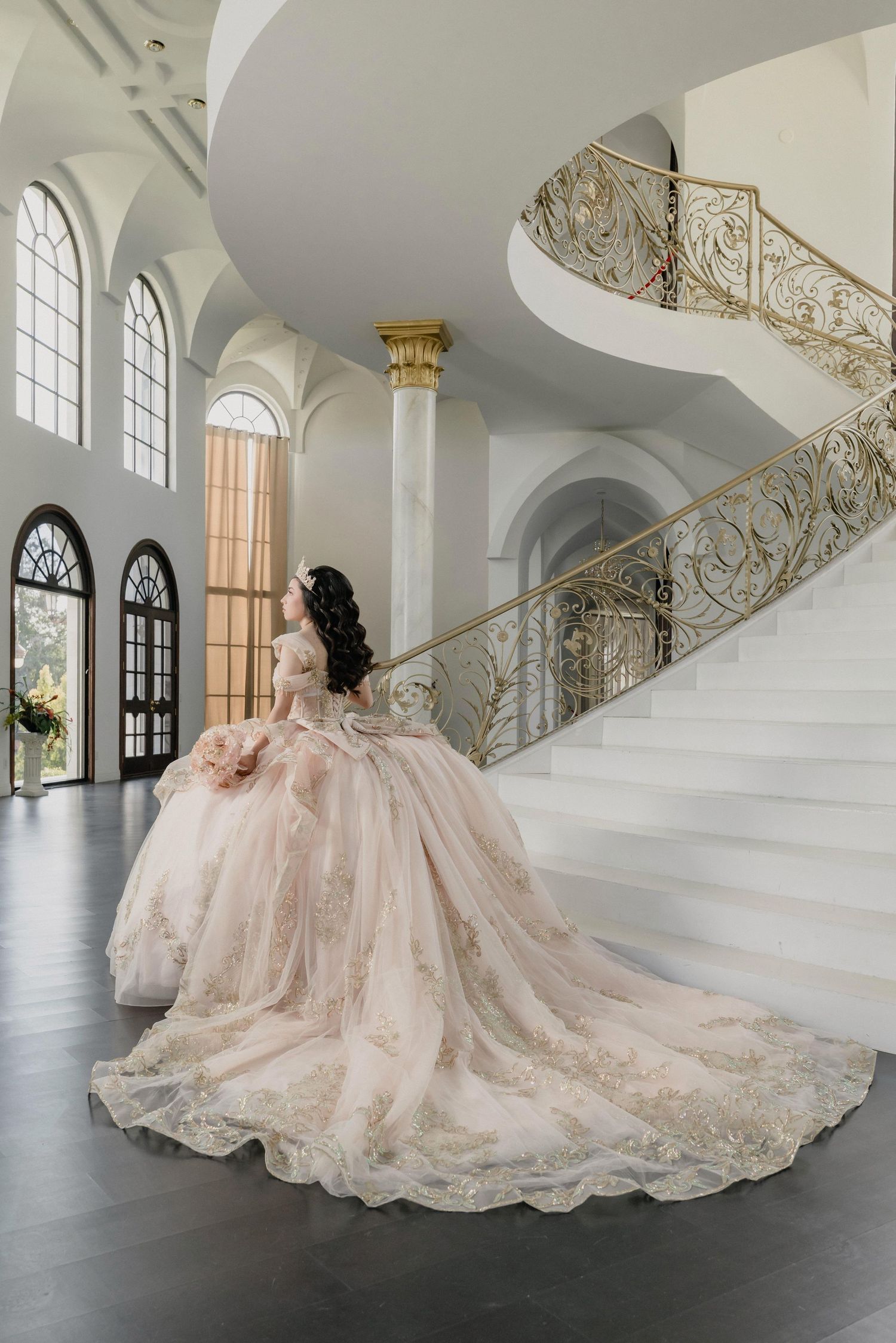 Elegant blush pink ballgown with intricate lace details flows across a grand white marble staircase in a luxurious setting.