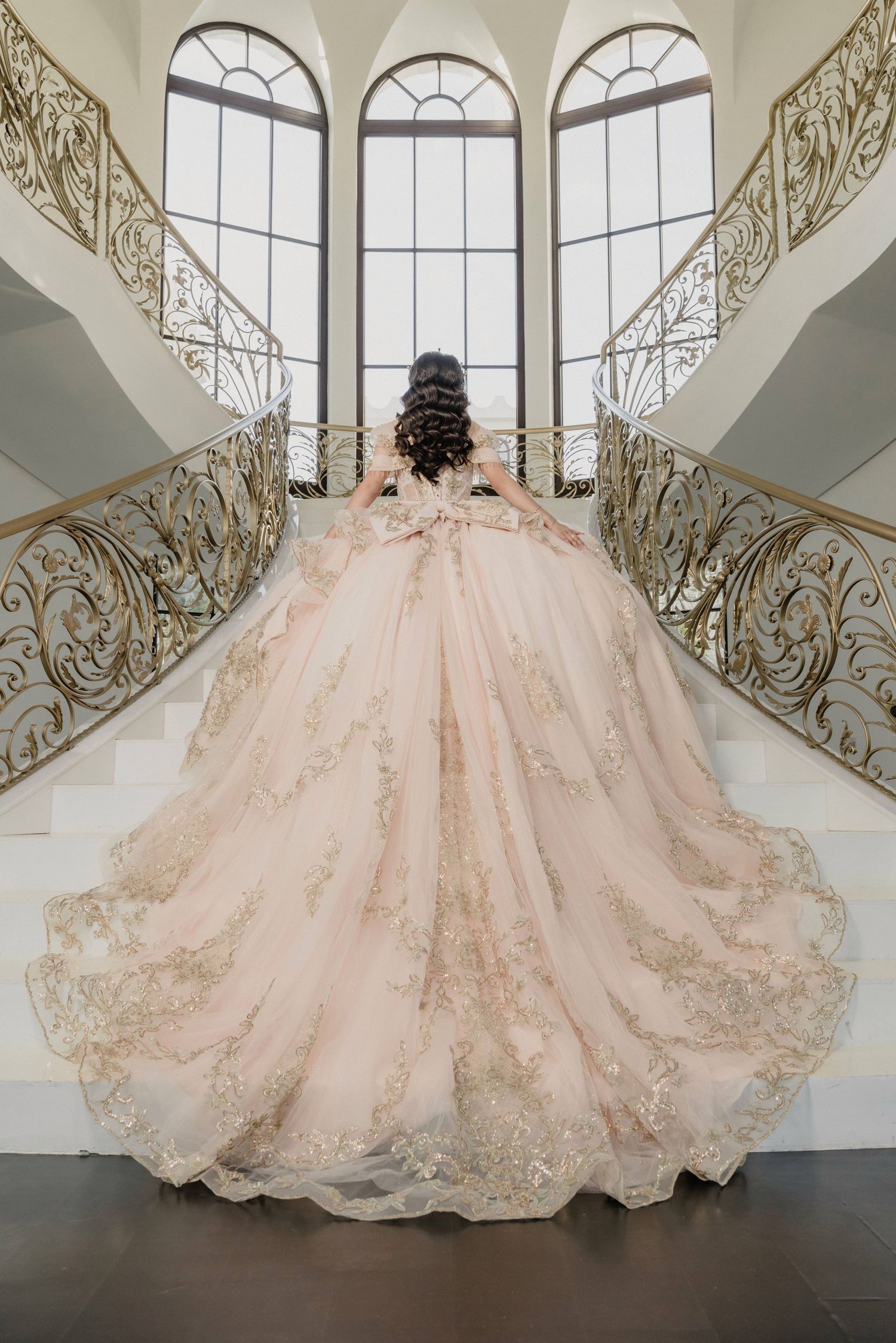 Luxurious blush pink ballgown with intricate lace details spreads across marble floor in elegant venue with arched windows.