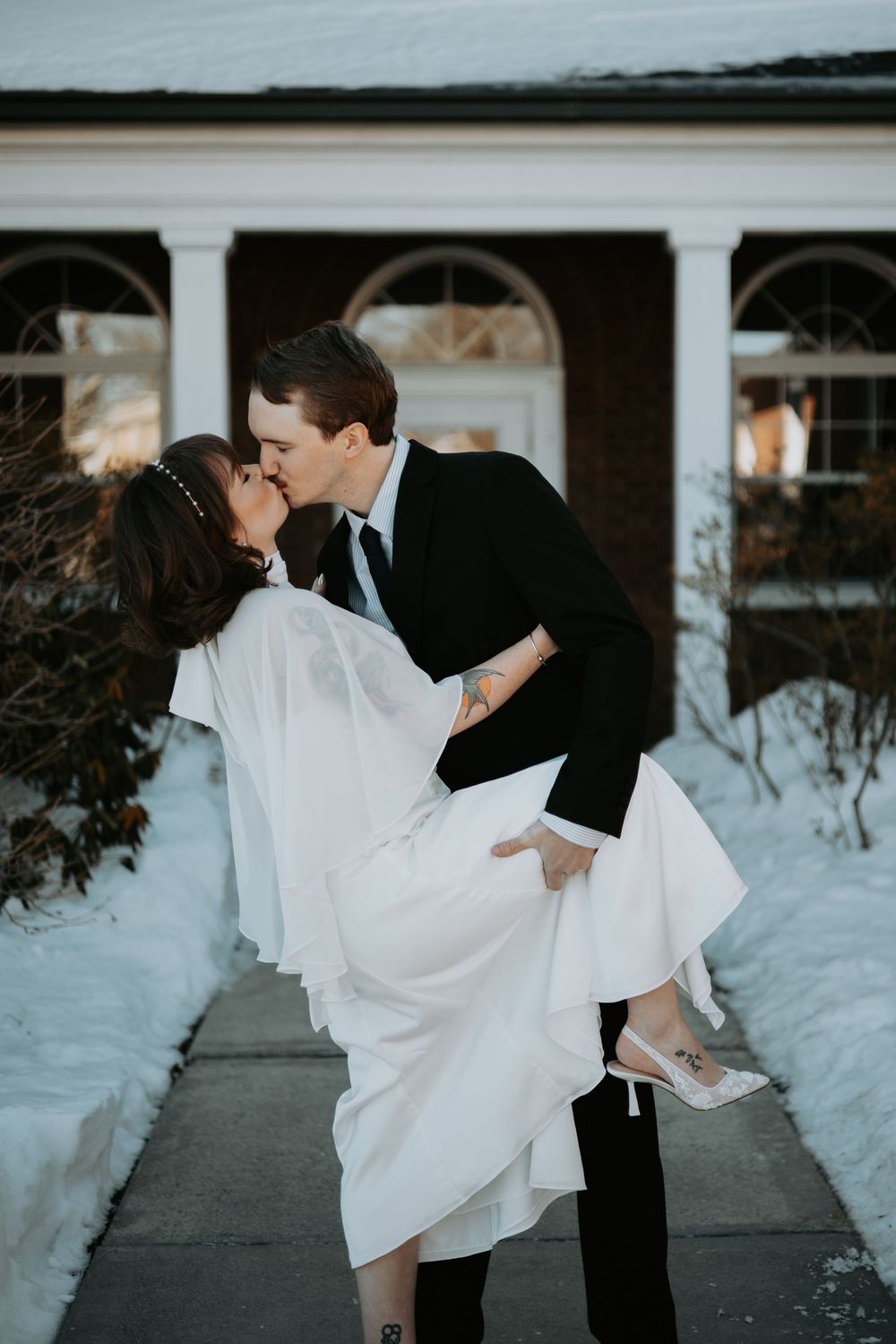 A romantic winter wedding couple shares intimate dance poses in the snow outside a building with arched windows.