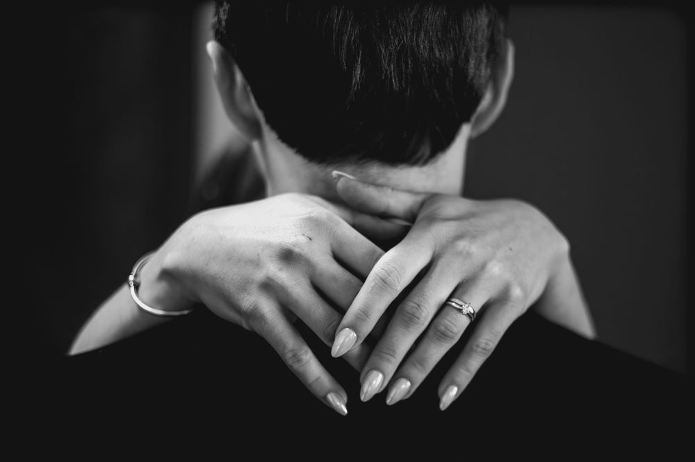 Black and white close-up of hands clasped behind a neck, with a wedding ring visible.