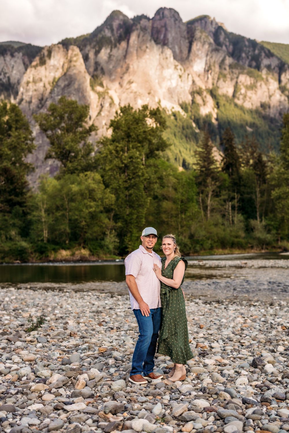 A couple shares a romantic moment on a rocky riverbed with towering mountains in the distance.