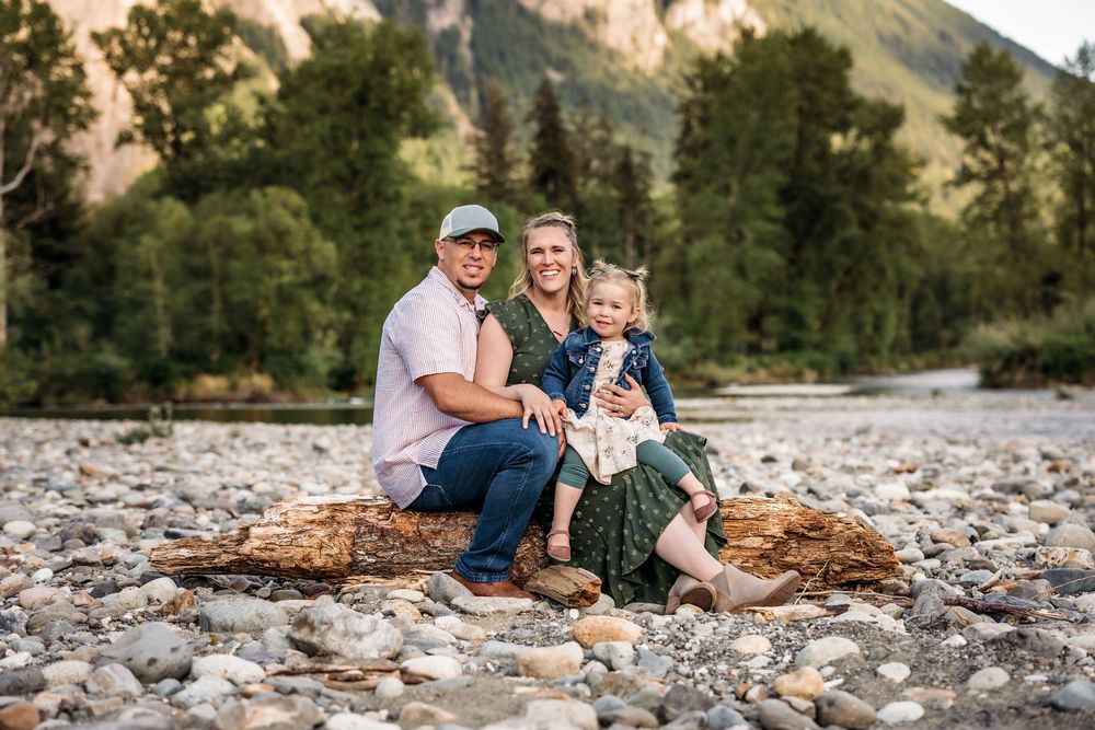 Family sitting together on rocky riverbank with mountain cliffs in background.