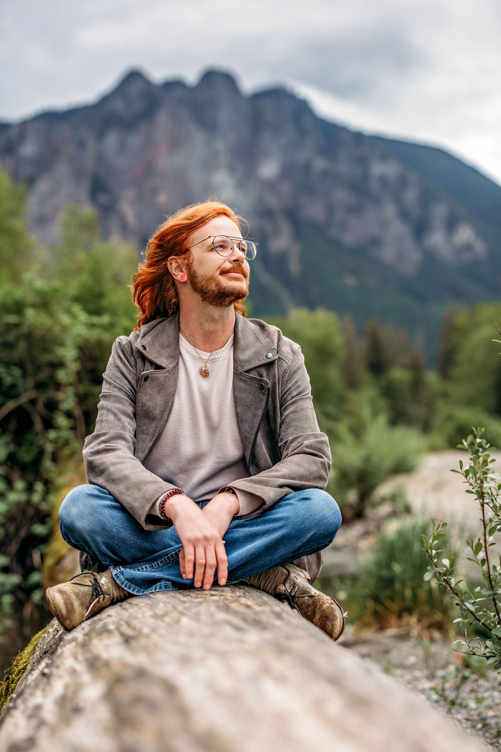 Person with red hair and glasses sits casually on rocks with mountain vista behind them.