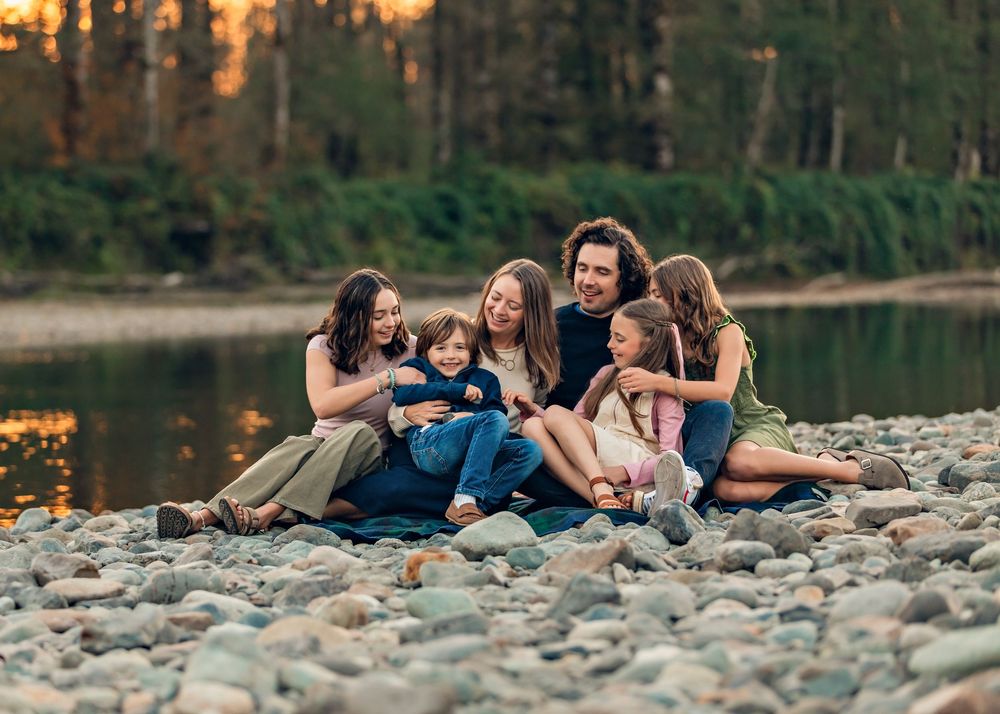 Group relaxing on rocky riverbank with mountains and forest in background during golden hour.