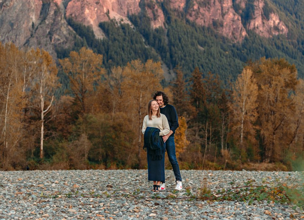 Family walking along rocky riverbed with mountain cliffs and autumn trees in background.