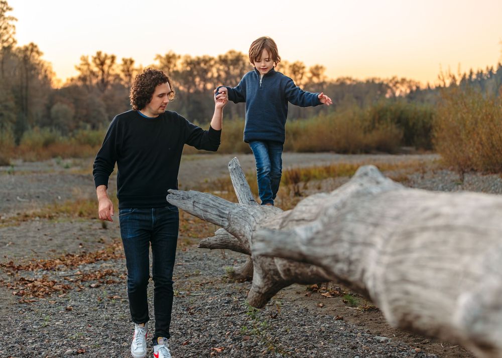 People walking and playing on driftwood logs at sunset by the river.
