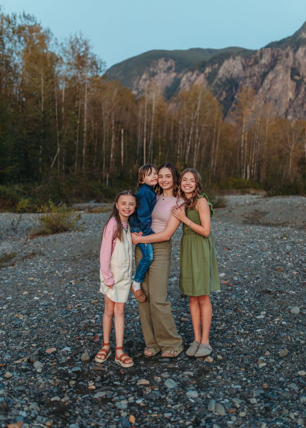 Group standing together on rocky riverbank with mountains and forest in background.