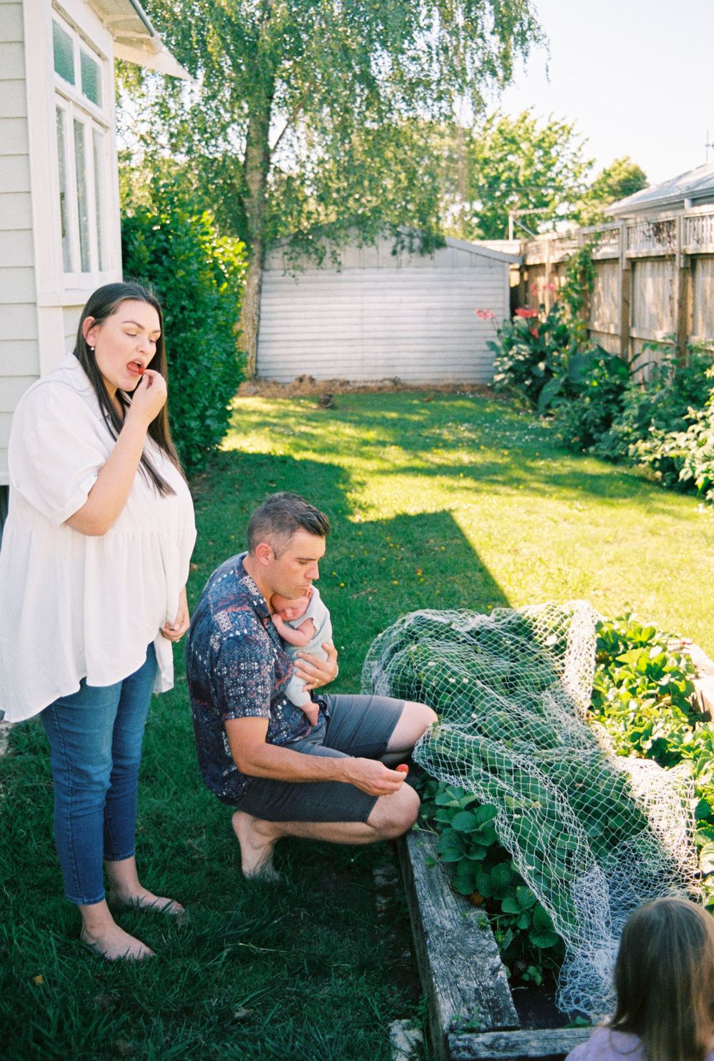 People share a light-hearted moment in a sunny backyard garden with lush greenery and white fencing.