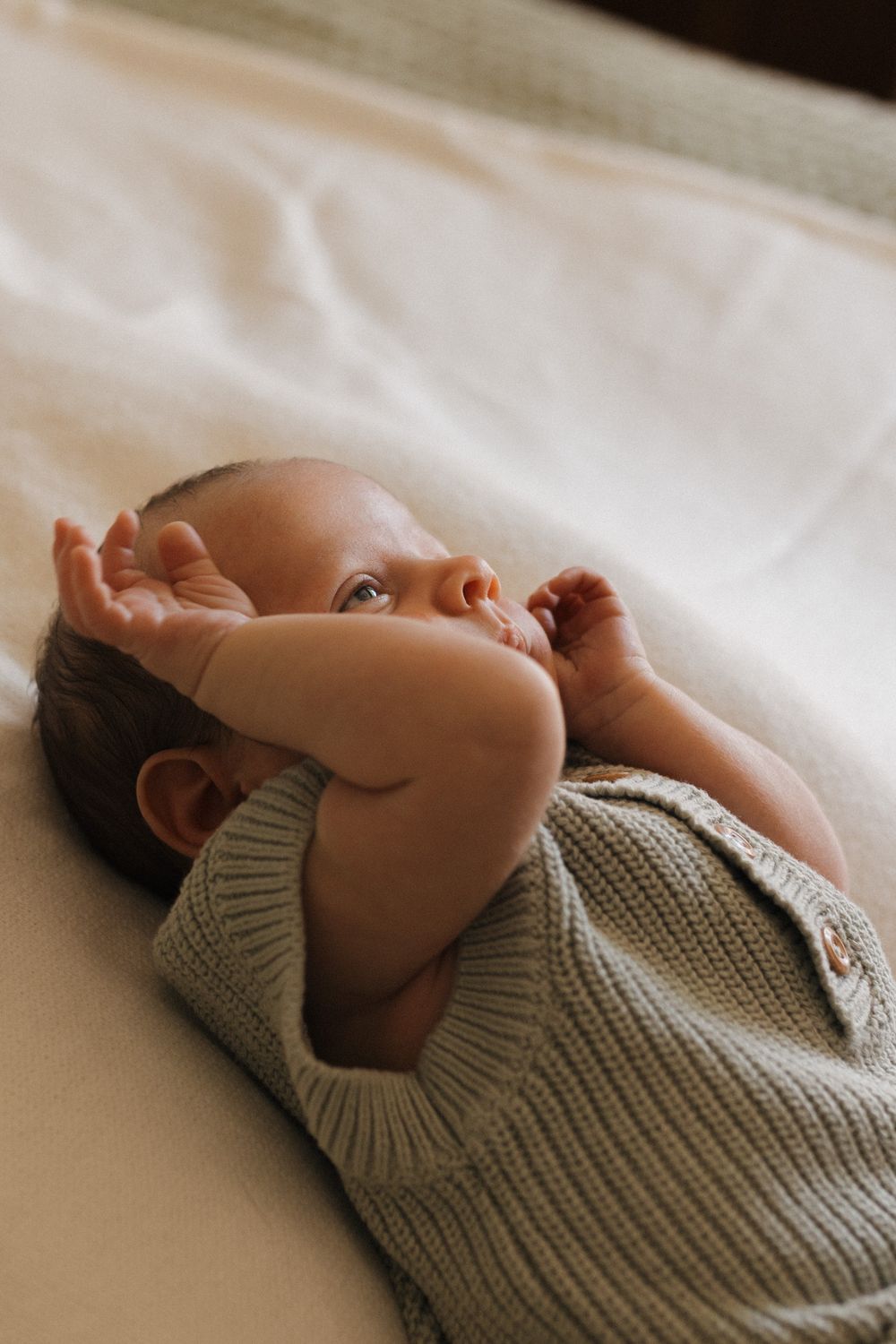 A peaceful portrait of a baby relaxing on soft white bedding wearing a cozy gray knit sweater.