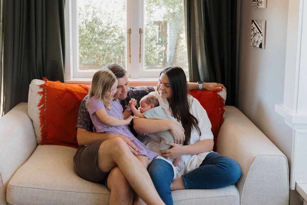 Family relaxing together on a white couch in a bright living room with orange accent pillows.