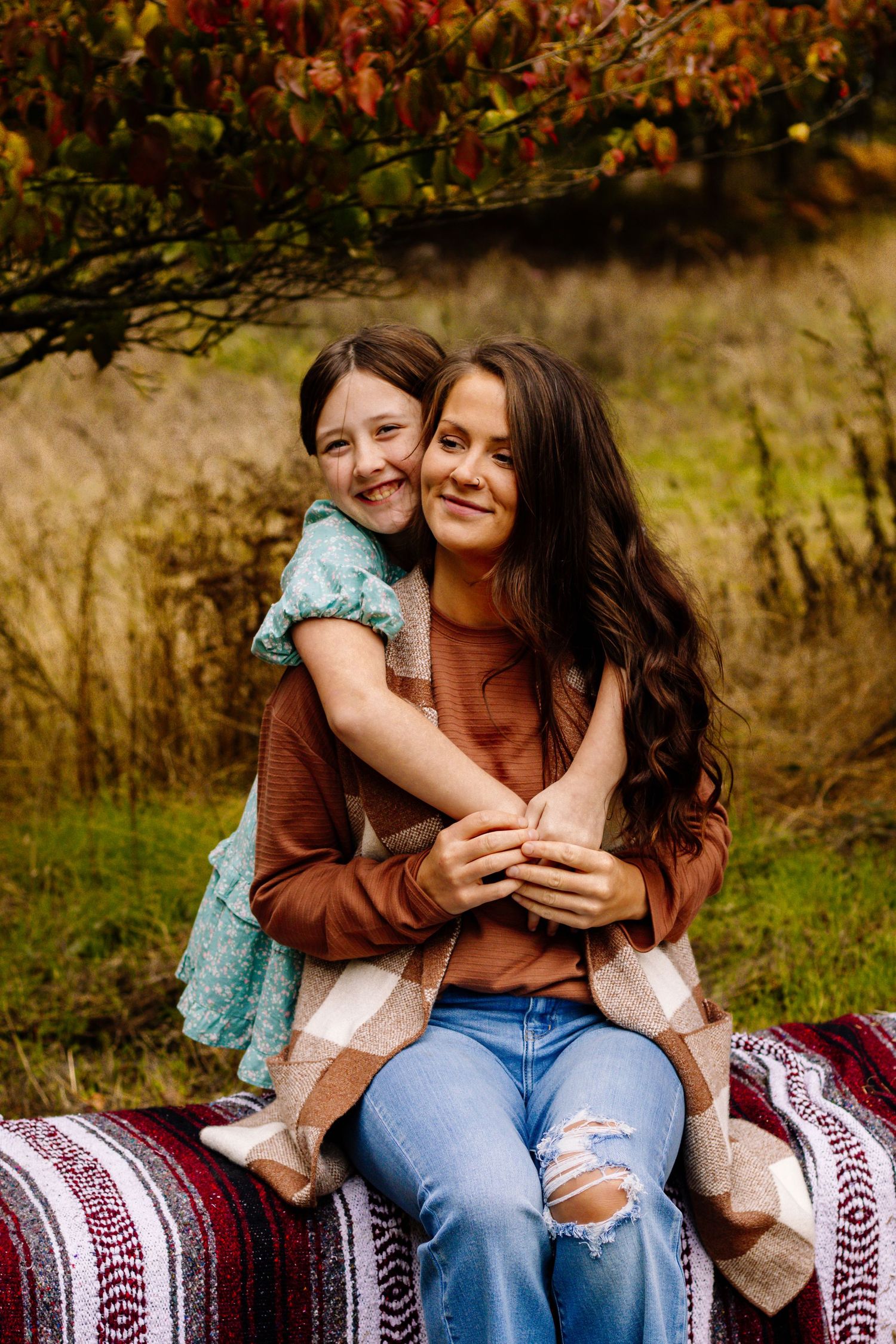 A mother and daughter share a warm embrace outdoors during autumn, sitting on a colorful blanket surrounded by fall foliage.