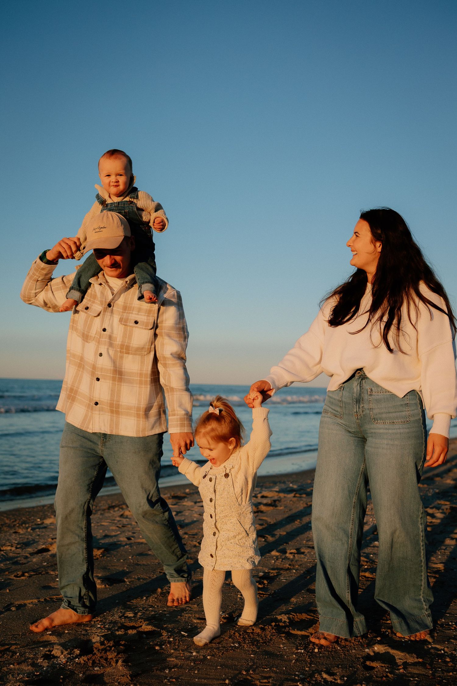 Family dressed in white plays together on a beach at sunset creating candid moments in warm golden light.
