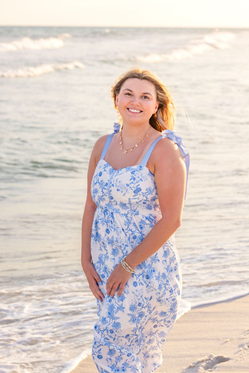 Woman in blue and white floral sundress poses on a sunny beach at sunset during a seaside photoshoot.