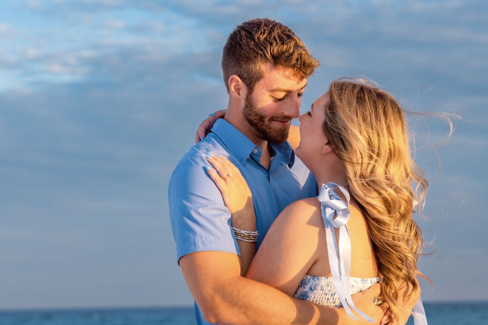 Two people embrace during a beach photoshoot with the ocean and soft blue sky creating a romantic backdrop.