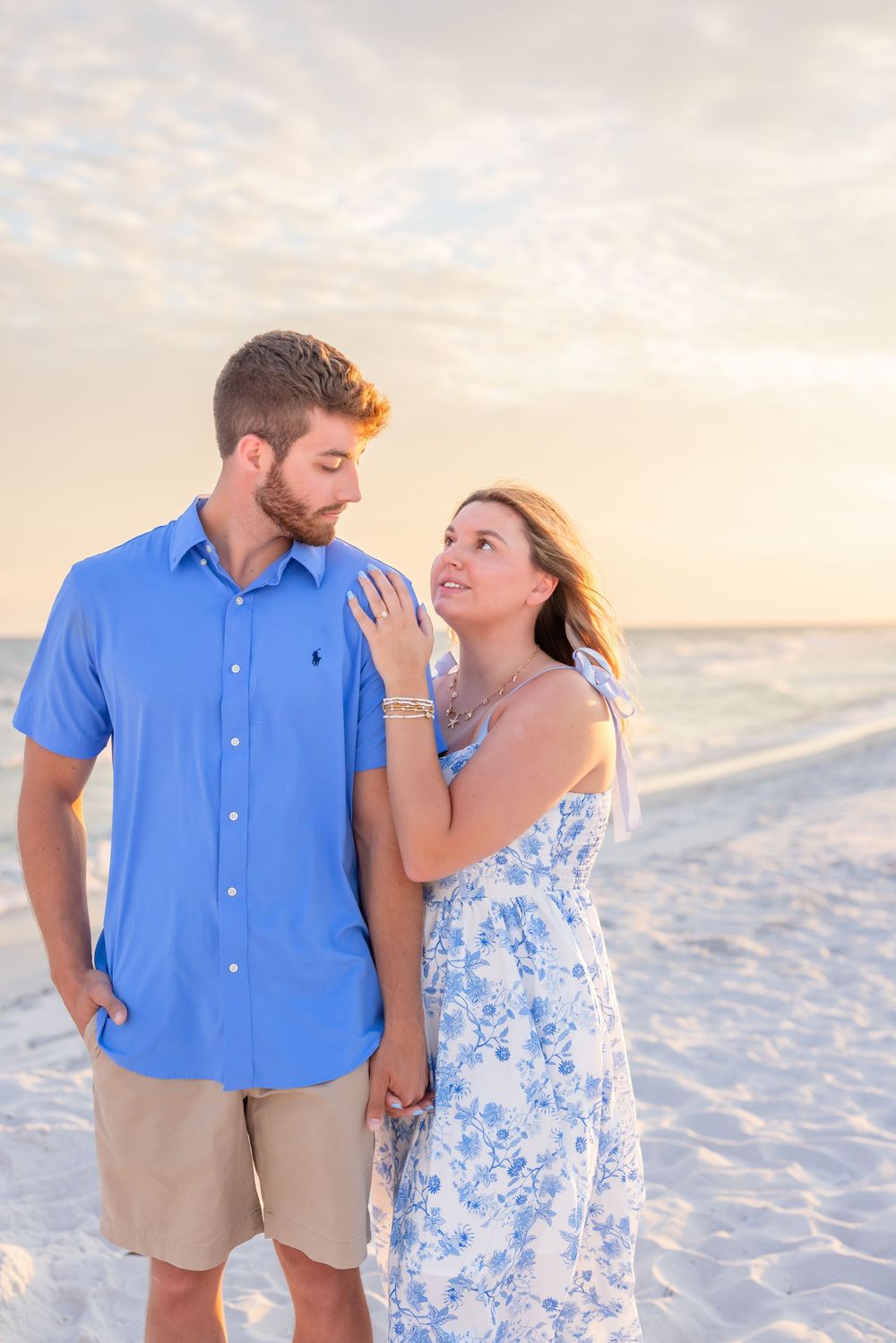 Couple wearing coordinating blue and white outfits embracing lovingly on a sunny beach at sunset.