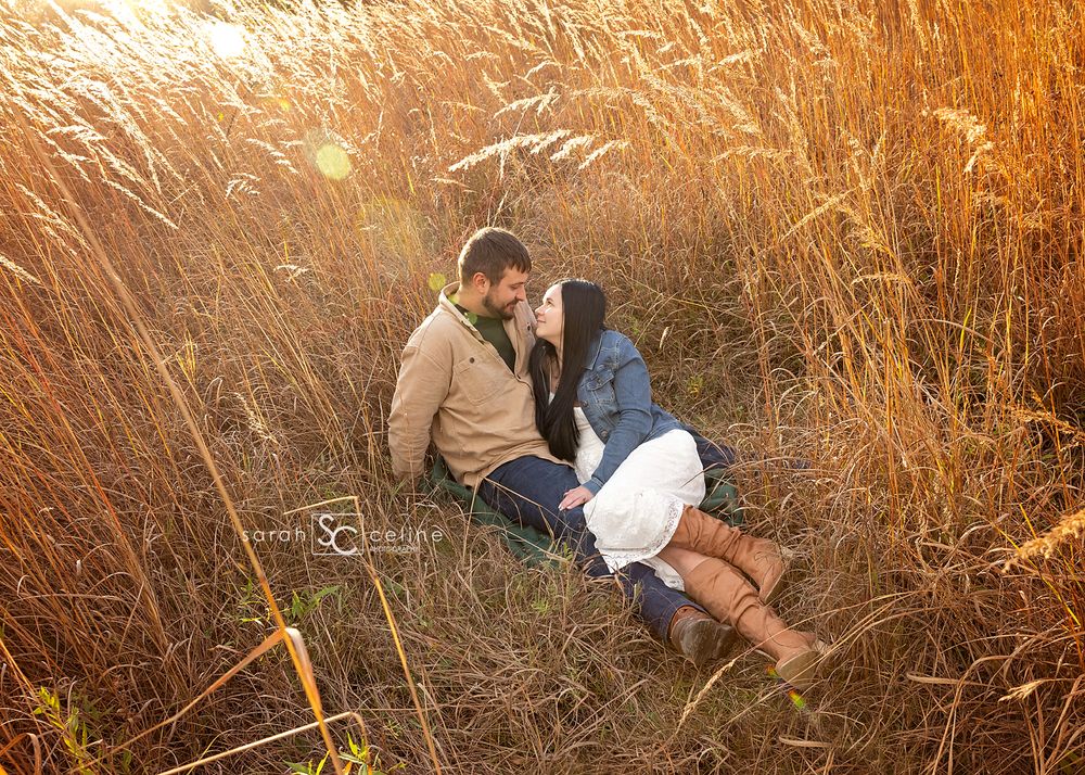 Couple sitting close together in tall golden grass at sunset, sharing an intimate moment during an outdoor engagement photo session.