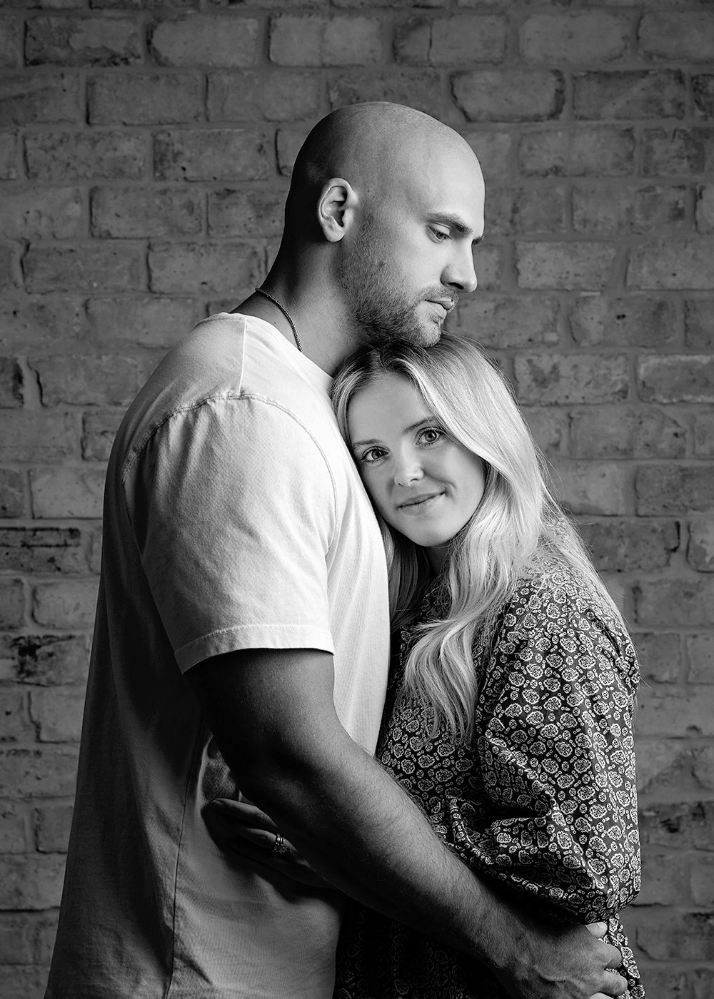 Black and white portrait of a couple embracing tenderly against a brick wall background.
