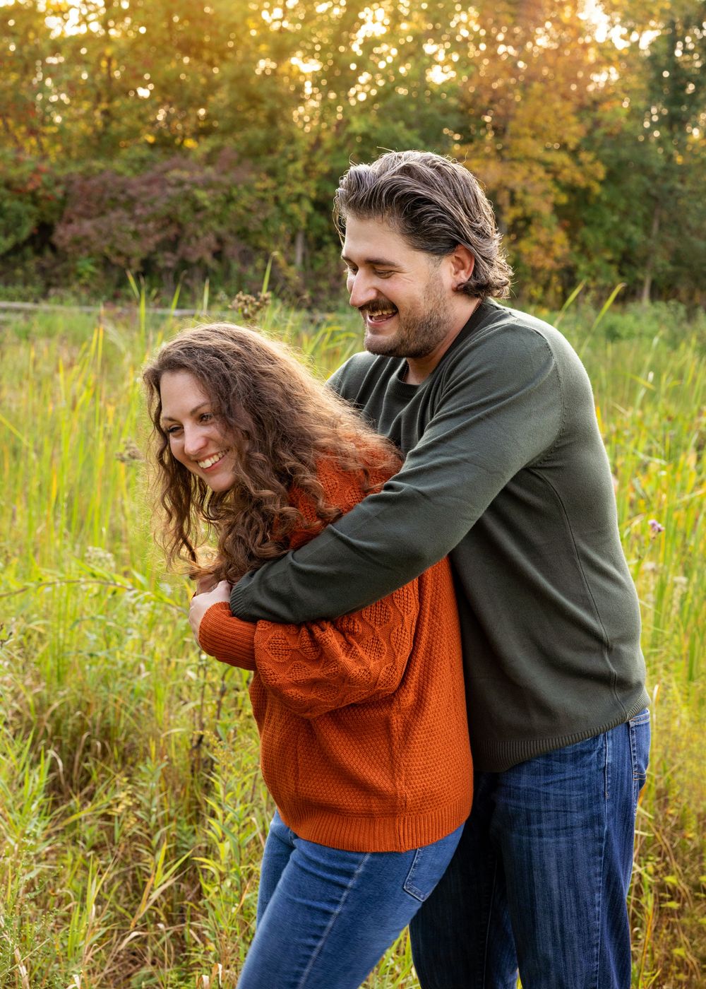 A couple embraces playfully in a golden autumn field during sunset, wearing orange sweater and dark jacket.